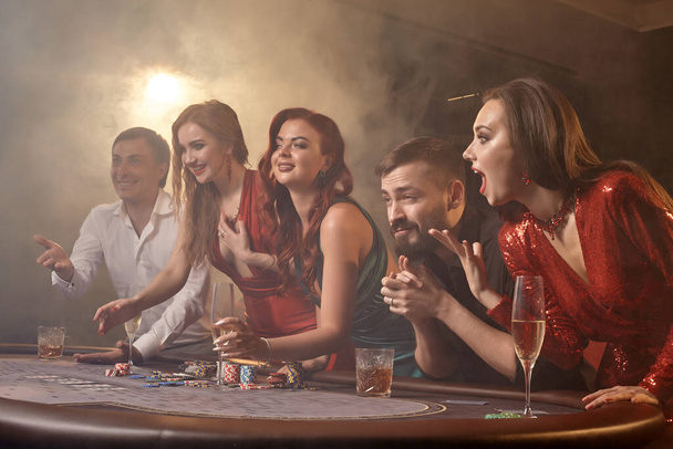 Excited buddies are playing poker at casino. Youth are making bets waiting for a big win while posing at the table against a white spotlight on a dark smoke background. Cards, chips, money, alcohol, gambling, entertainment concept. - Photo, image