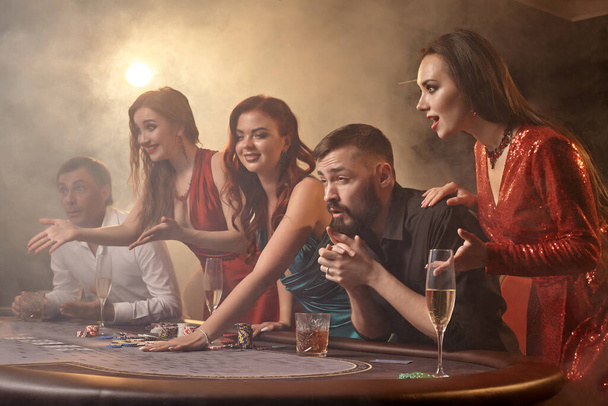 陽気な同僚はカジノでポーカーをしています。暗い煙の背景に白いスポットライトに対してテーブルにポーズをとっている間、若者は大きな勝利を待って賭けをしています。カード、チップ、お金、アルコール、ギャンブル、エンターテイメントの概念. - 写真・画像