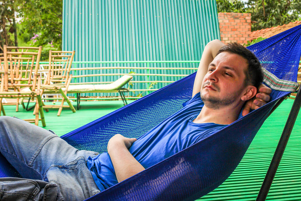 European guy dressed in a blue T-shirt and denim shorts is lying and resting in a blue hammock on the bamboo veranda of the hut, Vietnam. - Fotografie, Obrázek