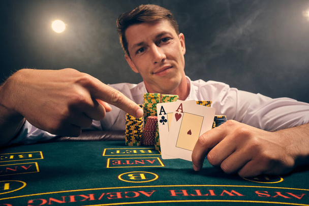 Detailní záběr chytrý chlapík v bílé košili hraje poker sedí u stolu v kasinu v kouři, proti bílému reflektoru. Radoval se z vítězství a ukazoval na dvě esa v ruce. Závislost na hazardu. Upřímné emoce a zábava co - Fotografie, Obrázek