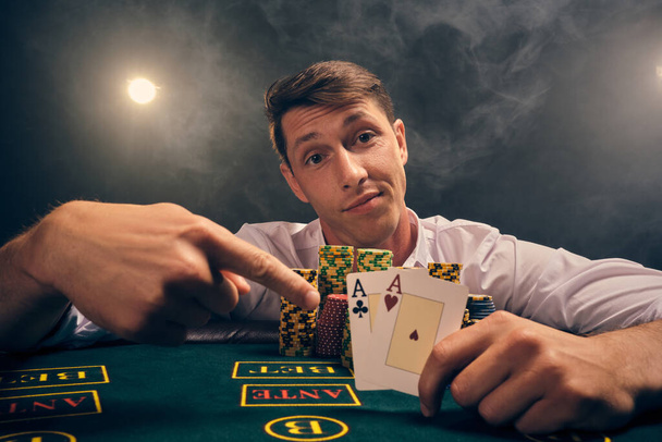Close-up shot of a smart person in a white shirt is playing poker sitting at the table at casino in smoke, against a white spotlight. He rejoicing his victory showing two aces in his hand. Gambling addiction. Sincere emotions and entertainment concep - Photo, Image