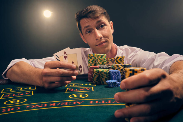 Close-up foto van een slimme man in een wit shirt is het spelen van poker zitten aan de tafel bij casino in rook, tegen een witte schijnwerper. Hij verheugt zich over zijn overwinning met twee azen en wat chips in zijn handen. Gokverslaving. Oprechte emoties en verdraagzaamheid - Foto, afbeelding