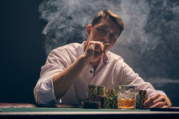 Handsome person in a white shirt is playing poker sitting at the table at casino in smoke, against a white spotlight. He rejoicing his victory and smoking a cigar. Gambling addiction. Sincere emotions and entertainment concept. - 写真・画像