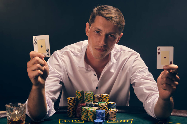 Handsome fellow in a white shirt is playing poker sitting at the table at casino in smoke, against a white spotlight. He rejoicing his victory showing two aces in his hands. Gambling addiction. Sincere emotions and entertainment concept. - Foto, afbeelding