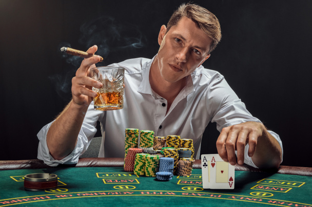 Un hombre majestuoso con una camisa blanca está jugando al póquer sentado en la mesa en el casino en humo, contra un foco blanco. Se regocijó por su victoria fumando un cigarro y bebiendo whisky, mostrando dos ases en su mano. Apuesta adicción. Emociones sinceras y en
 - Foto, Imagen