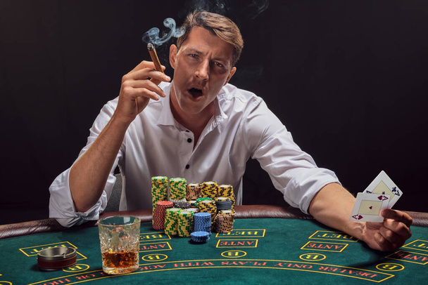 Komea kaveri valkoisessa paidassa pelaa pokeria kasinon pöydässä savuna ilmaan. Hän riemuitsee voitostaan ja polttaa sikaria samalla kun hänellä on kaksi ässää kädessään. Uhkapeliriippuvuus. Vilpittömät tunteet ja viihde käsite
. - Valokuva, kuva