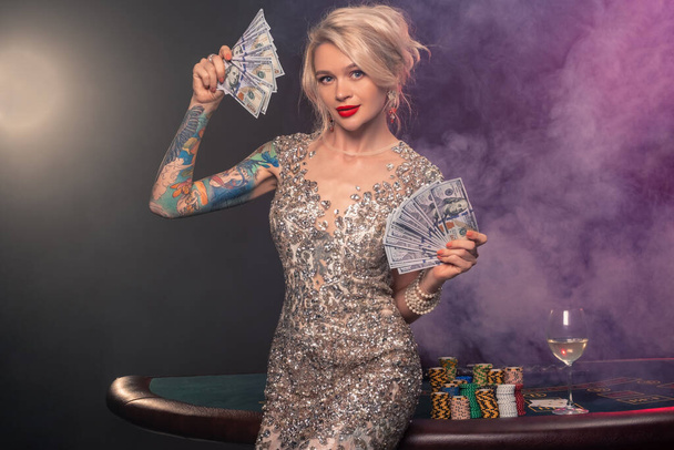 Medium close-up shot of a gorgeous girl with blond hair, tattoed hands and perfect make-up, dressed in a silver shiny dress. She is standing against a gambling table, with a fan of hundred dollar bills in her hands and smiling. Poker concept on a bla - Foto, Bild