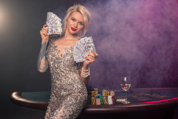 Medium close-up shot of a gorgeous maiden with blond hair, tattoed hands and perfect make-up, dressed in a silver shiny dress. She is standing against a gambling table, with a fan of hundred dollar bills in her hands and looking at you. Poker concept - Photo, Image