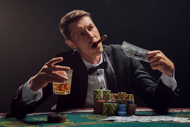 Όμορφος άντρας με μαύρο σλάζικ κοστούμι και λευκό πουκάμισο παίζει πόκερ στο τραπέζι του καζίνο σε καπνό, ενάντια σε ένα λευκό προβολέα. Χαιρόταν τη νίκη του καπνίζοντας ένα πούρο, πίνοντας αλκοόλ και κρατώντας κάποια χρήματα στο χέρι του. Τυχερά παιχνίδια a - Φωτογραφία, εικόνα