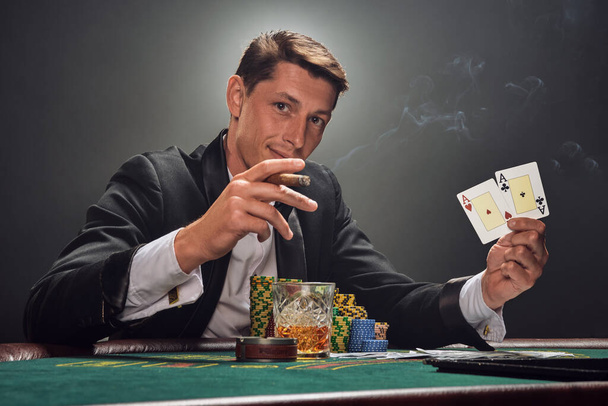 Hombre majestuoso en un traje negro slassic y camisa blanca está jugando al póquer sentado en la mesa en el casino en el humo, contra un foco blanco. Se regocijó con su victoria mostrando dos ases en su mano, fumando un cigarro y sonriendo a la cámara. Apuestas adicción
 - Foto, imagen