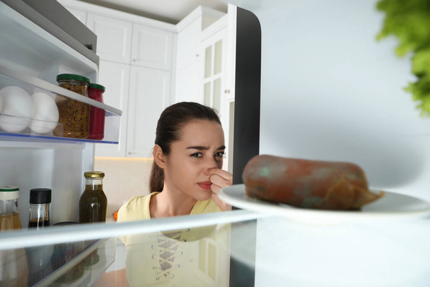 Young woman feeling bad smell of spoiled sausage in refrigerator, view from inside - Photo, Image