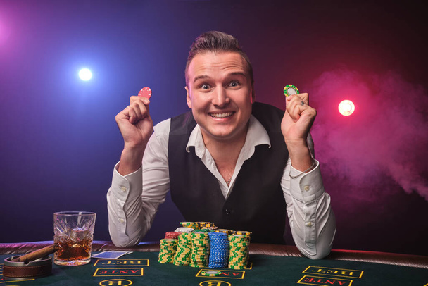 Πλούσιος ώριμος άντρας με λευκό πουκάμισο και μαύρο γιλέκο κρατάει μερικές μάρκες στα χέρια του και χαμογελάει. Παίζει πόκερ με ενθουσιασμό, ενώ κάθεται στο τραπέζι σε ένα καζίνο σε μια ακτίνα κόκκινων και μπλε backlights σε μαύρο φόντο. Επικίνδυνος τζόγος. - Φωτογραφία, εικόνα