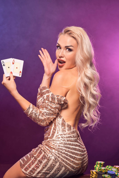 Nahaufnahme einer sexy Frau mit blonden Haaren und hellem Make-up, gekleidet in einem goldgestreiften, glänzenden Kleid, die mit zwei Assen in den Händen posiert, während sie auf einem Tisch mit Chips darauf sitzt und verwundert schaut. Poker-Konzept auf schwarzem Rauch-Hintergrund - Foto, Bild