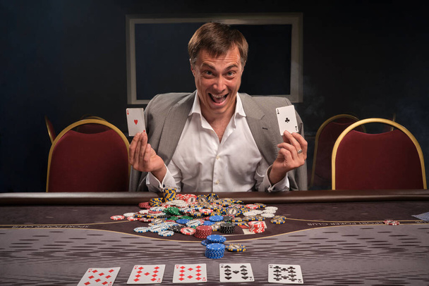 Joyful blond man in a white shirt and a light jacket is playing poker sitting at the table in casino. He rejoices in winning holding two aces in his hands. Gambling for money. Games of fortune. - Photo, Image