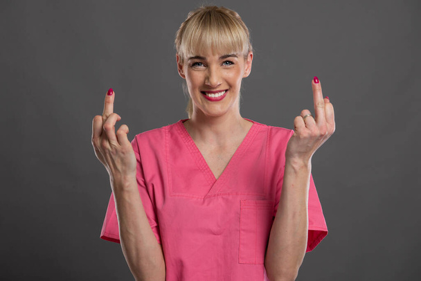 Retrato de una joven enfermera atractiva mostrando doble dedo medio sobre fondo gris
 - Foto, Imagen