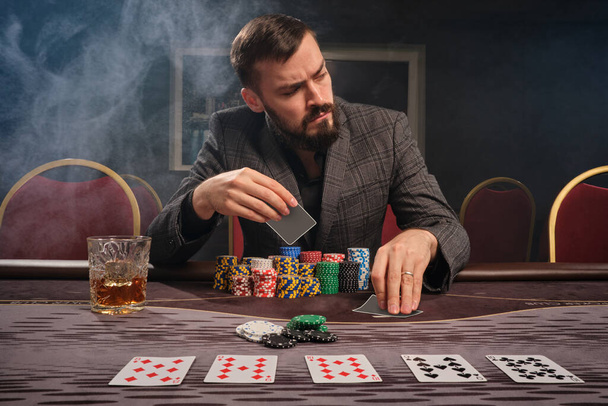 Ο γενειοφόρος τύπος με το γκρι κλασικό κοστούμι, με ένα ποτήρι ουίσκι παίζει πόκερ καθισμένος στο τραπέζι στο καζίνο. Βάζει στοιχήματα περιμένοντας μια μεγάλη νίκη. Τζόγος για λεφτά. Παιχνίδια τύχης. - Φωτογραφία, εικόνα