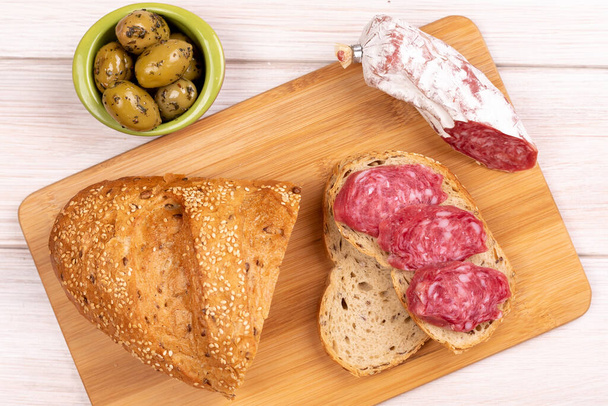 Сэндвич из сушеных сосисок, оливок и буханки цельного зернового хлеба на деревянной доске. Концепция простой доступной еды. Вид сверху
 - Фото, изображение