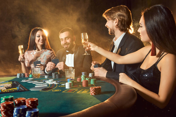 Freunde der Oberschicht spielen in einem Casino. Zwei Männer in Anzügen und zwei junge Frauen in Kleidern. Rauch. Kasino. Poker - Foto, Bild