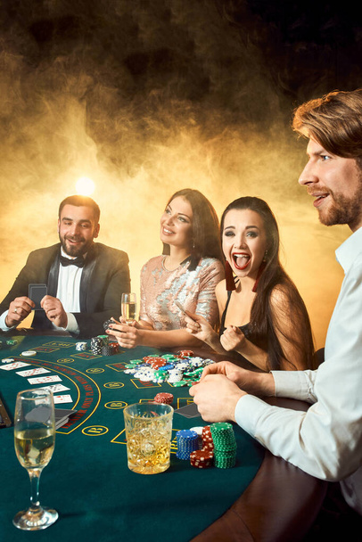 Přátelé z vyšší třídy hrají v kasinu. Dva muži v oblecích a dvě mladé ženy v šatech. Kouř. Kasino. Poker - Fotografie, Obrázek