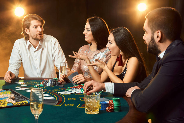 Amici di classe superiore che giocano d'azzardo in un casinò. Due uomini in giacca e cravatta e due giovani donne in abito. Fumo. Casinò. Poker
 - Foto, immagini