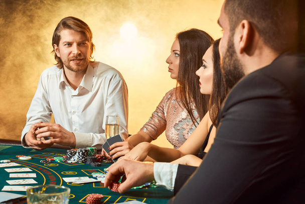Yläluokan ystävät uhkapeli kasinolla. Kaksi miestä puvuissa ja kaksi nuorta naista mekoissa. Savua. Kasino. Pokeri
 - Valokuva, kuva