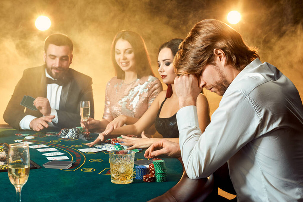 Φίλοι της ανώτερης τάξης τζογάρουν σε καζίνο. Δύο άντρες με κοστούμια και δύο νεαρές γυναίκες με φορέματα. Καπνός. Καζίνο. Πόκερ - Φωτογραφία, εικόνα
