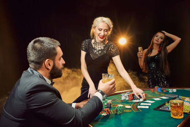 Eine Gruppe junger reicher Leute pokert im Casino. Mann im Businessanzug und zwei junge Frauen in schwarzen Kleidern. Rauch. Kasino. Poker - Foto, Bild