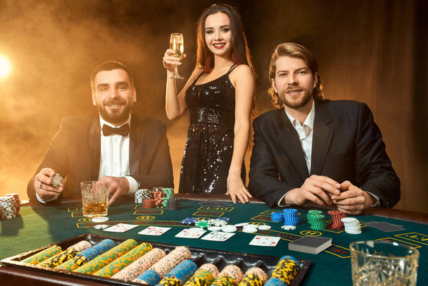 Fiatal, gazdag emberek csoportja pókerezik a kaszinóban. Két öltönyös férfi és egy fekete ruhás fiatal nő. Füst. Kaszinó. Póker - Fotó, kép