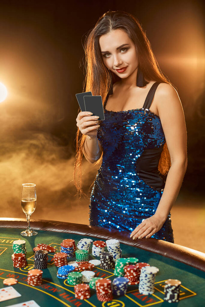 Jovem mulher bonita em um vestido azul brilhante posa perto da mesa de poker no casino de luxo. Uma jogadora. Paixão, cartões, batata frita, álcool, dados, jogo, cassino - é como entretenimento feminino. Perigoso divertido jogo de cartas por dinheiro. Fundo de fumo
. - Foto, Imagem