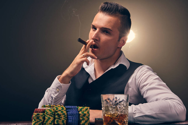 Serious mature person in a white shirt and black waistcoat is smoking a cigar and looking at the camera. He is playing poker at a casino in a ray of backlight on black background. Risky gambling entertainment with cards and chips. - Fotografie, Obrázek