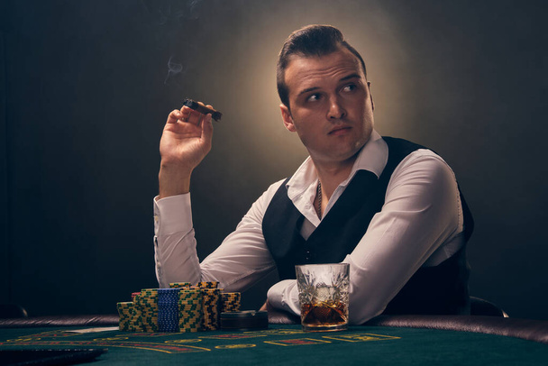 Rijke volwassen man in een wit shirt en zwarte vest houdt een sigaar in zijn hand en kijkt weg. Hij speelt poker in een casino in een straal van achtergrondverlichting op zwarte achtergrond. Riskant gokken entertainment met kaarten en chips. - Foto, afbeelding