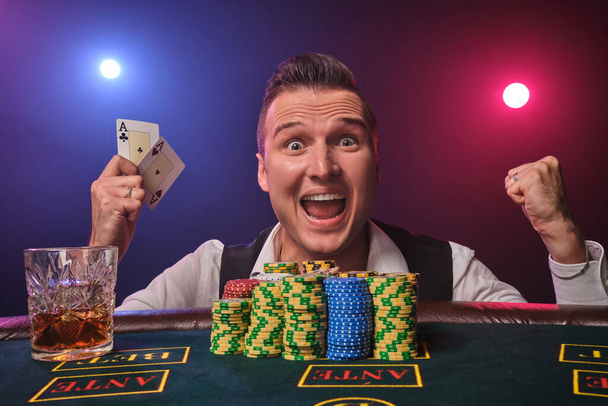 A sportos, érett férfi fehér ingben és fekete mellényben két kártyajátékot tart a kezében. Egy pókerasztalnál pózol zsetonokkal és egy pohár whiskyvel egy kaszinóban, piros és kék háttérvilágítással fekete háttérben. Kockázatos szerencsejáték - Fotó, kép