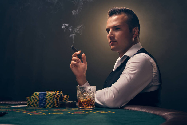 Όμορφος ώριμος άντρας με λευκό πουκάμισο και μαύρο γιλέκο καπνίζει πούρο κάθεται πλάγια στο τραπέζι και κοιτάζει αλλού. Παίζει πόκερ σε ένα καζίνο σε μια αχτίδα φωτός σε μαύρο φόντο. Επικίνδυνη ψυχαγωγία τυχερών παιχνιδιών με κάρτες και μάρκες - Φωτογραφία, εικόνα