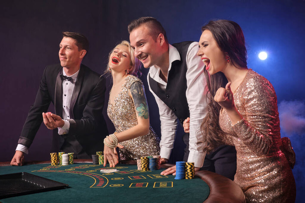 Side shot of a joyful rich companions playing poker at casino in smoke. Youth are making bets waiting for a big win. They are looking excited standing at the table against a red and blue backlights on black background. Risky gambling entertainment. - Photo, Image
