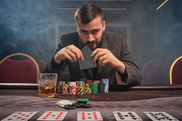 Bearded guy in a gray suit is playing poker sitting at the table at casino in a smoke. He is holding two empty cards in his hand and looking at the camera. Gambling addiction. Entertainment concept. - Foto, Imagem