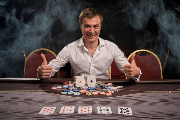 Захоплений усміхнений хлопець у білій сорочці грає в покер, сидячи за столом в казино в димі. Він радіє перемозі, показуючи великі пальці вгору і посміхаючись. Азартна залежність. Концепція розваг
. - Фото, зображення