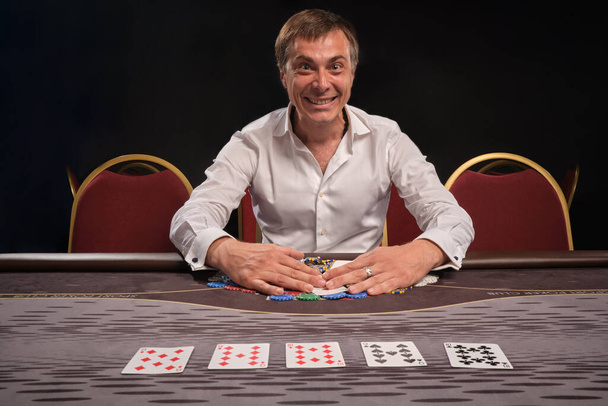 白いシャツを着た興奮したカリスマ男性がカジノのテーブルに座ってポーカーをプレイしています。彼は勝利を喜び、チップを持っている。ギャンブル中毒。エンターテインメントコンセプト. - 写真・画像