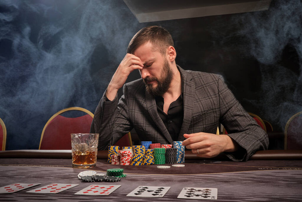 Bearded guy in a gray suit is playing poker sitting at the table at casino in a smoke. He closed his eyes and looking upset. Gambling addiction. Entertainment concept. - Fotografie, Obrázek