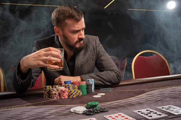 Bearded man in a gray suit is playing poker sitting at the table at casino in a smoke. He is holding a glass of whiskey in his hand and looking away. Gambling addiction. Entertainment concept. - Photo, Image