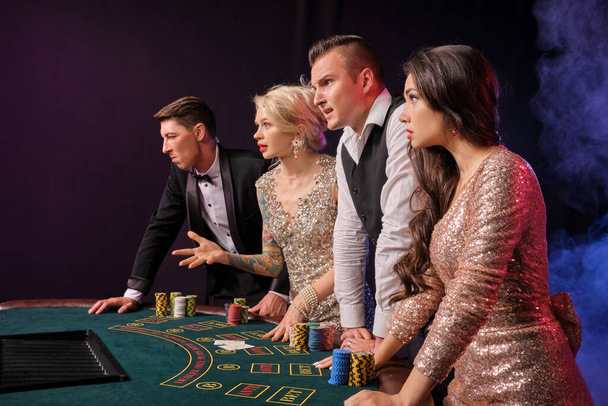 Side shot of a disappointed rich buddies playing poker at casino in smoke. Youth have lose. They are looking unhappy standing at the table against a red and blue backlights on black background. Risky gambling entertainment. - Photo, Image