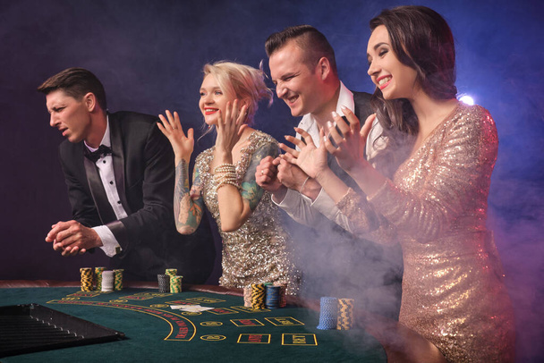 Zdjęcie boczne bogatych kumpli grających w pokera w kasynie z dymem. Młodzież robi zakłady czekając na wielką wygraną. Klaszczą dłońmi stojącymi przy stole na czerwono-niebieskim podświetleniu na czarnym tle. Ryzykowne rozrywki hazardowe - Zdjęcie, obraz