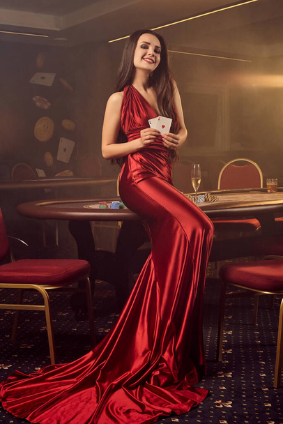 Charming brunette girl in a long burgundy atlas dress, with two aces in her hands is smiling and posing sideways near a poker table in vip casino. Passion, cards, chips, alcohol drinks, win, gambling - it is a best entertainment. Smoke background. - Foto, Imagen