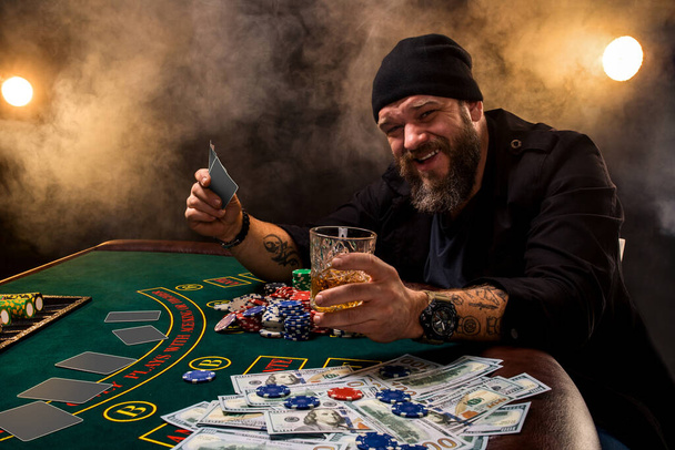 Szakállas férfi szivarral és üveggel a pókerasztalnál egy kaszinóban. Szerencsejáték, kártyázás és rulett. A zöld pókerasztalon kártyák, zsetonok és pénz van. Az egész szoba füstöl a szivaroktól. - Fotó, kép