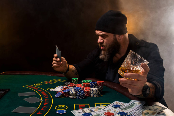 Szakállas férfi szivarral és üveggel a pókerasztalnál egy kaszinóban. Szerencsejáték, kártyázás és rulett. A zöld pókerasztalon kártyák, zsetonok és pénz van. Az egész szoba füstöl a szivaroktól. - Fotó, kép