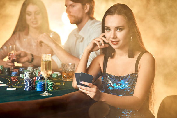 Group of an elegant people playing poker at the gambling house. Focus on a luxurious brunette in a blue shiny dress. Passion, cards, chips, alcohol, dice, gambling, casino - it is entertainment. Dangerous fun card game for money. Smoke background. - Photo, Image