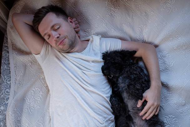 Junger Kaukasier, der im Bett schläft und seinen Hund umarmt. Bleiben Sie zu Hause. Ansicht von oben - Foto, Bild