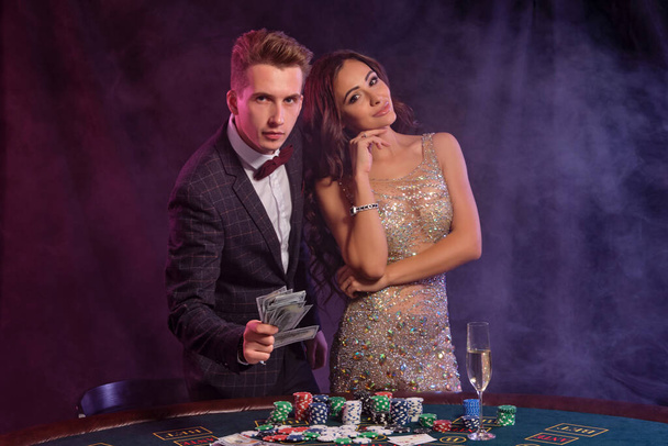 Man and woman are showing cash, playing poker at casino and celebrating their win at table with stacks of chips, money, cards, champagne on it. Black, smoke background, colorful backlights. Close-up. - Photo, Image
