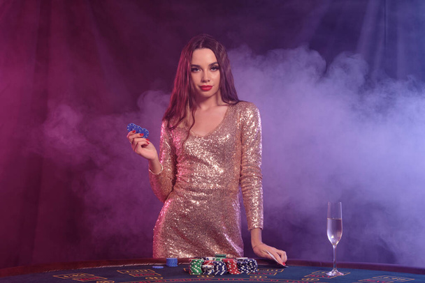 Kaunis nainen kultainen mekko näyttää kaksi pelimerkkiä poseeratessaan pöydässä kasinolla. Musta, savutausta, värikkäät taustavalot, samppanja. Uhkapeliviihdettä, pokeria. lähikuva
. - Valokuva, kuva
