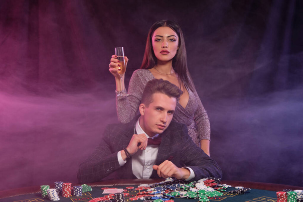 カジノでポーカーをする男は、チップ、お金、シャンパン、カードの山とテーブルに座っています。女性との勝利を祝う。黒の背景。閉鎖. - 写真・画像