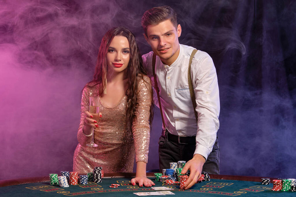 Мужчина и женщина играют в покер в казино, празднуют победу за столом с пачками фишек, карт, шампанского. Черный, дымовой фон. Крупный план
. - Фото, изображение